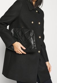 Bolso de mano acolchado negro con un acabado brillante, que presenta un patrón geométrico y un cierre dorado, sostenido contra un abrigo negro de doble botonadura.