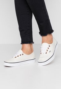 Weiße Leinenschuhe mit kupferfarbenen Ösen, weißen Schnürsenkeln und einem schwarzen Streifen entlang der Sohle. Getragen mit schwarzen ausgefransten Jeans.