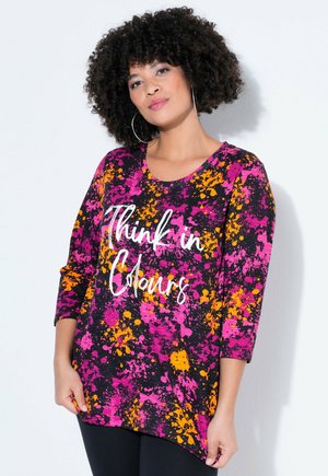 Mujer con cabello rizado que lleva grandes pendientes de aro y una blusa colorida en tonos rosa, naranja y negro con el texto "Think in Colours".