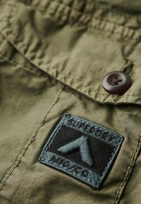 Tela verde oliva con un acabado texturizado, que presenta un parche azul oscuro con el texto "SUPERDRY" y un diseño de triángulo negro, asegurado por un botón marrón.