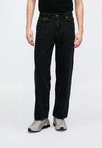 Jeans de ganga preto de perna straight com costura proeminente, cinco bolsos e fecho de botão. Usados com sapatilhas de desporto cinza claro.