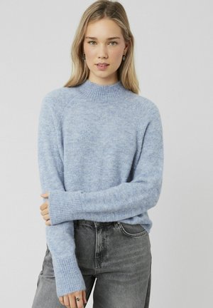 Pull bleu clair en tissu doux et texturé avec un col montant côtelé et des manches longues. Associé à un jean gris foncé.