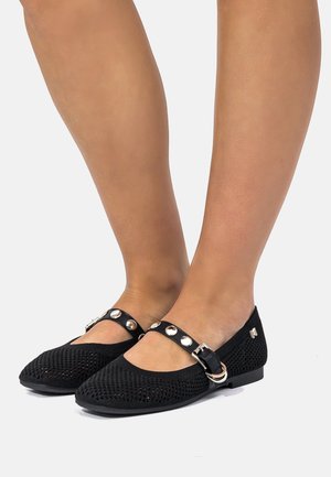 Pies con zapatos tipo Mary Jane de punto negro con correa tachonada y hebilla sobre un fondo gris claro.