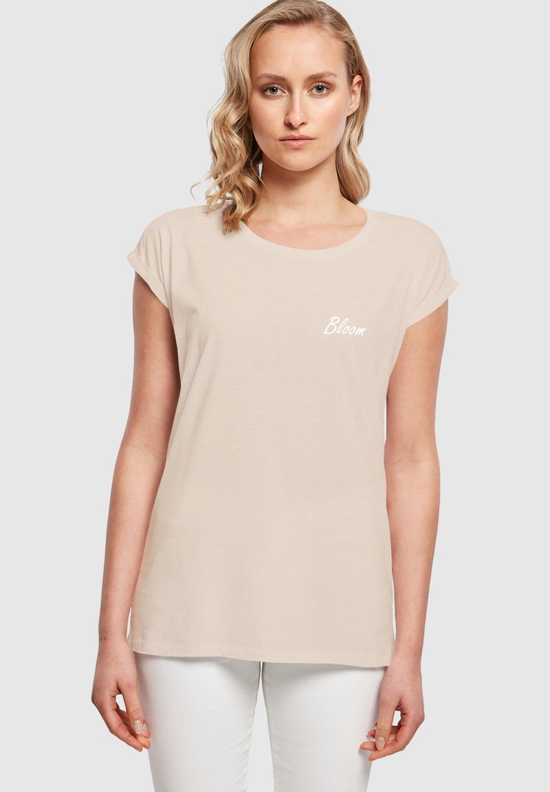 Beige puuvillane T-särk lühikeste rulli varrukatega, ümar kaelus ja ees valge "Bloom" tekst. Kandsid valgeid pükse.