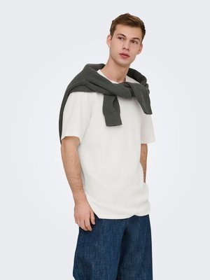 Jeune homme regardant sur le côté, portant une chemise blanche à manches courtes, un pantalon bleu foncé, avec un pull gris foncé posé sur ses épaules.