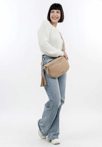 Bolso cruzado de piel beige con cierre de cremallera, con un acento de borlas y correa ajustable. Se lleva un suéter blanco de punto y jeans azules.