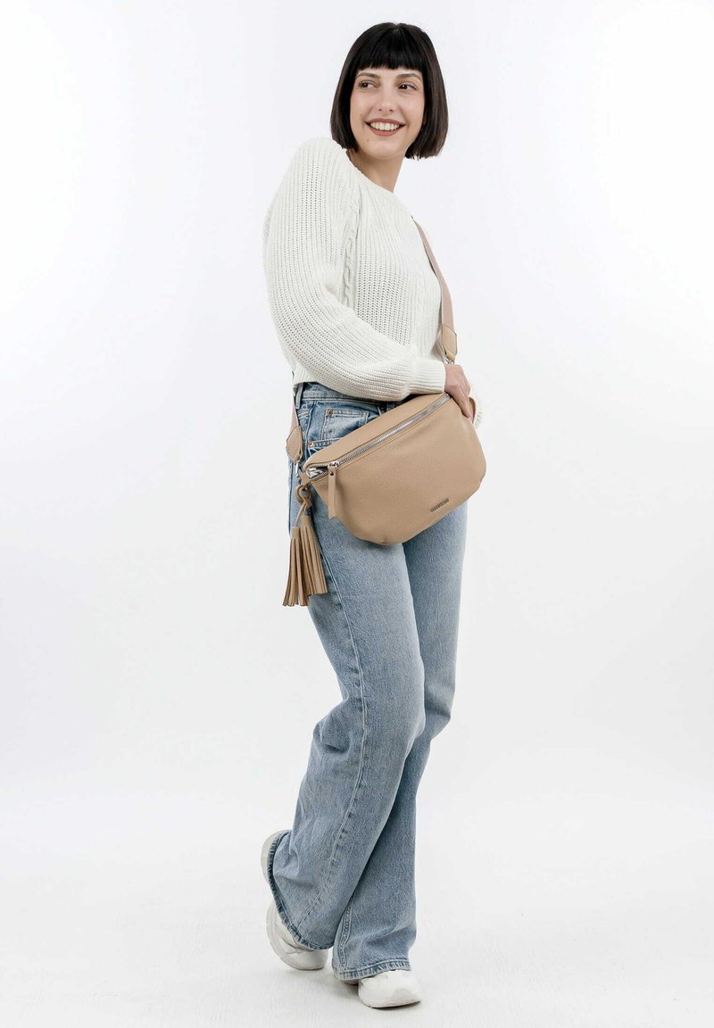 Bolso cruzado de piel beige con cierre de cremallera, con un acento de borlas y correa ajustable. Se lleva un suéter blanco de punto y jeans azules.