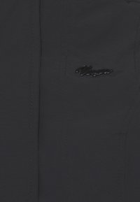 Dunkelgraue, leichte Jacke mit glatter Textur, auffälligen Nähten und einem glänzenden schwarzen Logo auf der linken Seite.