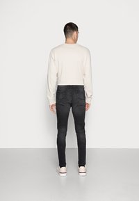 Top longo de manga comprida, bege claro, com decote redondo, combinado com jeans skinny pretos. Modelo usando ténis bege com detalhes em vermelho.