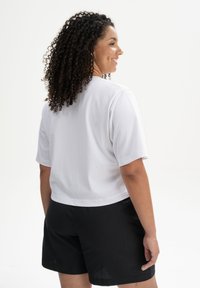 Weißes, kurz ärmeliges, cropped T-Shirt mit rundem Hals, kombiniert mit schwarzen Shorts. Glatte Textur, einfaches Design und keine sichtbaren Muster.