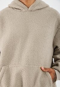 Bežs fleeces kapucis džemperis ar mīkstu, teksturētu virsmu, ar saņemamu kapuci, kangaru kabatu un kontrastējošas krāsas piedurknēm.