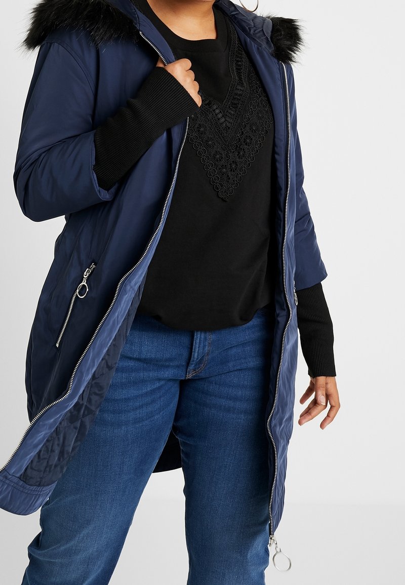 Granatowa pikowana kurtka z futrzanym obszyciem kaptura, zamkiem z przodu i bocznymi kieszeniami, noszona na czarnym swetrze z teksturowanym dekoltem.