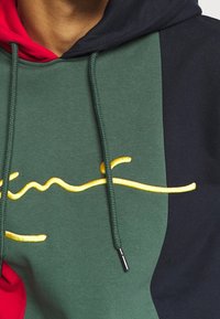 Felpa con cappuccio caratterizzata da un design a blocchi di colore in verde, rosso e blu navy. La parte frontale presenta testo in giallo scritto in corsivo e fibbie per i lacci testurizzate.