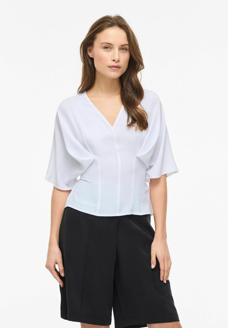 Femme aux longs cheveux bruns portant un haut blanc à col en V avec des manches larges et un short noir taille haute, debout devant un fond blanc.