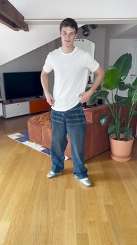 Osoba stoji u dnevnoj sobi, noseći bijelu majicu i plave traperice. Traperice imaju horizontalne šavove. Na nogama ima srebrne tenisice.