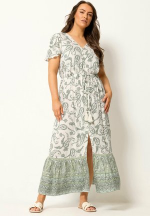 Femme portant une robe maxi blanche et verte à motif cachemire, manches courtes, boutonnée devant, ceinture à la taille et fente à l’avant, associée à des sandales blanches à enfiler.
