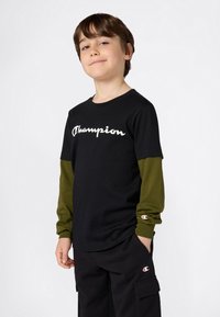 Kind, das ein schwarzes Champion-T-Shirt über einem grünen Langarmshirt und schwarzen Cargo-Hosen trägt, mit den Händen in den Taschen, steht selbstbewusst vor einem weißen Hintergrund.