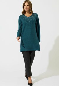 Tunique lavorata a maglia color verde acqua con scollatura a V, decorata con paillettes sul petto, abbinata a pantaloni aderenti neri e tacchi neri.