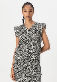 Svart-hvitt mønstrete bluse med V-hals og volangærmer. Stoffet er lett, med et geometrisk design og flytende silhuett.