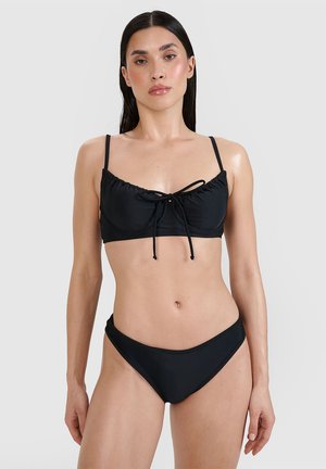 Kobieta o długich ciemnych włosach nosi czarne bikini z wiązanym detalem na górze, stoi na jednolitym jasnoszarym tle.