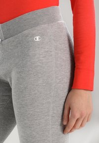 Graue Sweatpants mit geripptem Bund, mit weißen Logodetails und glatter Textur. Kombiniert mit einem roten Langarmshirt.