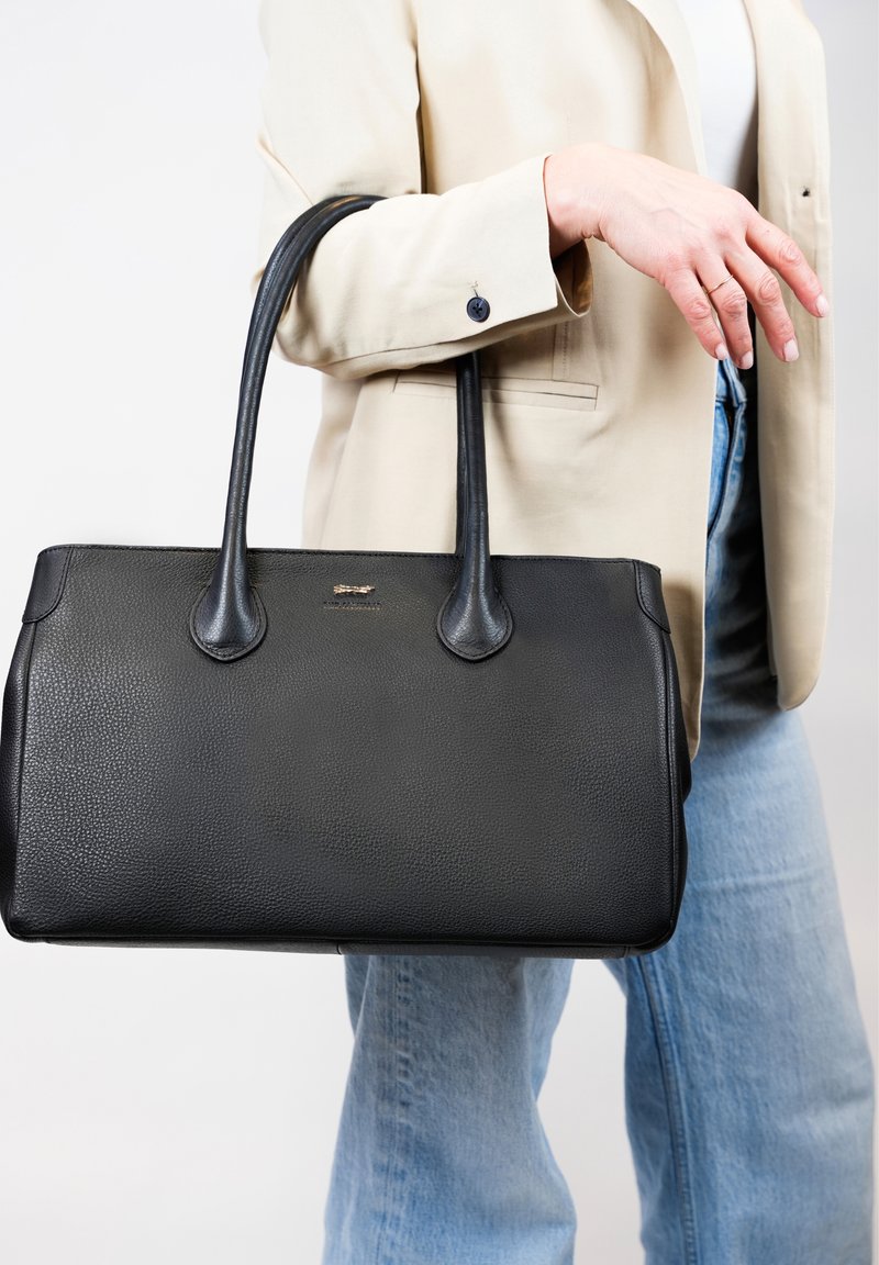 Zwarte leren tote bag met een gestructureerde afwerking, dubbele handgrepen en een gouden accentlogo. Gepresenteerd tegen een neutrale achtergrond.