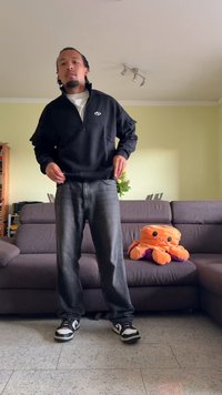 Μαύρη φούτερ με φερμουάρ, λευκό μπλουζάκι από κάτω, σκούρο γκρι τζιν και μαύρα παπούτσια. Καθισμένο σε έναν γκρι καναπέ βρίσκεται ένα πορτοκαλί μαλακό παιχνίδι όκτων.