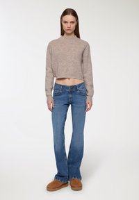 Młoda kobieta ubrana w beżowy sweter w krótkim fasonie, niebieskie dżinsy z wysokim stanem i podwójnymi guzikami, oraz brązowe zamszowe buty na platformach, stojąca na białym tle.