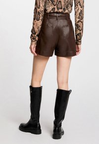Femme portant un short en cuir marron foncé, un haut à manches longues en imprimé serpent et des bottes noires jusqu'au genou, se tenant devant un fond uni.