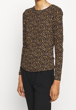 Personne portant un haut noir à manches longues avec un petit imprimé floral jaune et orange, associé à un pantalon noir sur fond blanc.