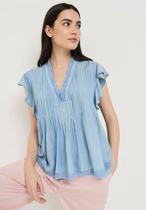 Femme portant un chemisier plissé bleu clair avec bordure en dentelle et manches volantées, associé à un pantalon rose clair, regardant sur le côté.