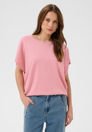 Femme aux longs cheveux bruns portant un haut rose ample à manches courtes, un jean bleu et un collier en argent, debout avec une main dans la poche.