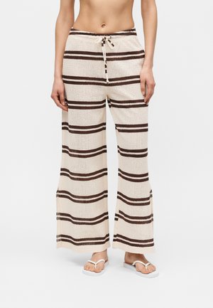 Frau trägt cremefarbene Strickhose mit horizontalen dunkelbraunen Streifen, weiten Beinen und einem Kordelzug in der Taille, kombiniert mit weißen Flip-Flops.