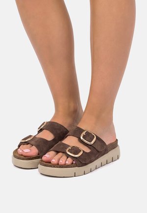 Piedi con unghie dei piedi dipinte di rosa chiaro che indossano sandali di camoscio marrone con due fibbie dorate regolabili e suole spesse beige.