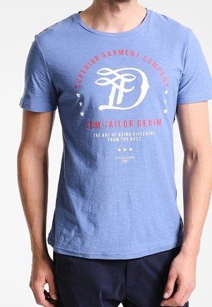 Blaues T-Shirt mit kurzen Ärmeln und weißem sowie rotem Text "Superior Garment Company Tom Tailor Denim", getragen von einer stehenden Person.