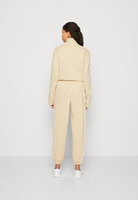 Light beige tracksuit with a high collar, ribbed cuffs, and elastic waistband. Features side stripes with letters and white sneakers.
