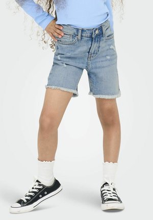 Shorts en jean avec ourlet effiloché, délavage clair, détails usés, associés à des baskets noires et des chaussettes blanches à bord ondulé.