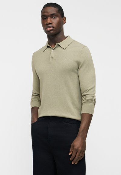 Homme portant un polo à manches longues beige clair avec deux boutons, pantalon noir, debout les mains dans les poches, sur un fond blanc.
