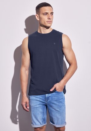 Marineblaue ärmellose Baumwollshirt mit runder Ausschnitt, kombiniert mit leicht blauen Jeansshorts mit aufgerollten Ärmeln. Minimalistisches Branding-Detail auf dem Shirt.