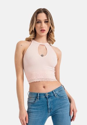 Femme portant un crop top côtelé rose pâle avec bordure en dentelle et une découpe en forme de goutte d'eau, assorti à un jean bleu taille mi-haute sur un fond blanc.
