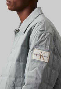 Hellegraue gesteppte Jacke mit Druckknopfverschluss und strukturierter Oberfläche. Mit einem Calvin Klein Jeans Logo-Patch auf dem linken Ärmel.