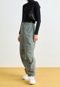 Kvinna klädd i svart kofta med dragkedja, olivgröna cargobyxor, vita sneakers och en liten svart axelväska, stående på gul golv.