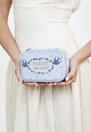 Ruky držia svetlomodrý obdĺžnikový vak s nápisom „FORGET ME NOT“, kvetinovým vencom a dvoma vyšívanými vtáčikmi na pozadí bielej sukne.