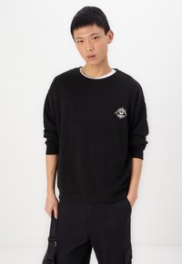Zwart crewneck sweatshirt met geribbelde manchetten, voorzien van een klein wit geborduurd zonnetje op de linkerkant van de borst. Zacht materiaal, casual pasvorm.