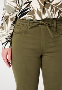 Femme portant un pantalon vert olive avec un taille élastique et un blouse à manches longues avec un motif abstrait de feuilles beige, noir et blanc.