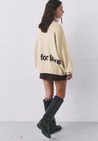 Pulover supradimensionat bej cu text "for love" în negru, realizat dintr-un tricot texturat. Purtați deasupra unei fuste închise și a unor cizme negre până la genunchi.