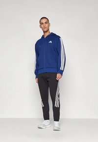 Casaco azul com fecho de correr e riscas brancas nas mangas, combinado com calças de treino pretas com riscas brancas nas laterais. Modelo em pose neutra.