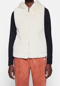 Krémová bezrukávová vesta s měkkou, chlupatou texturou a kapucí; má zipové zapínání a sahá po pás, nošena přes černý dlouhý rukáv.