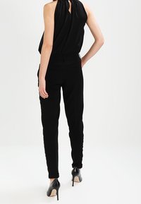 Svart ärmlös topp med draperad halsringning, tillsammans med figurnära svarta byxor. Outfitten har högklackade svarta skor.