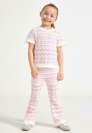 Hymyilevä tyttö seisoo, pukeutuneena vaaleanpuna-valkoiseen neulottuun lyhythihaiseen yläosaan ja samaa sarjaa oleviin housuihin, joissa on aaltoilevat reunat, harmaat lenkkarit valkoisella taustalla.